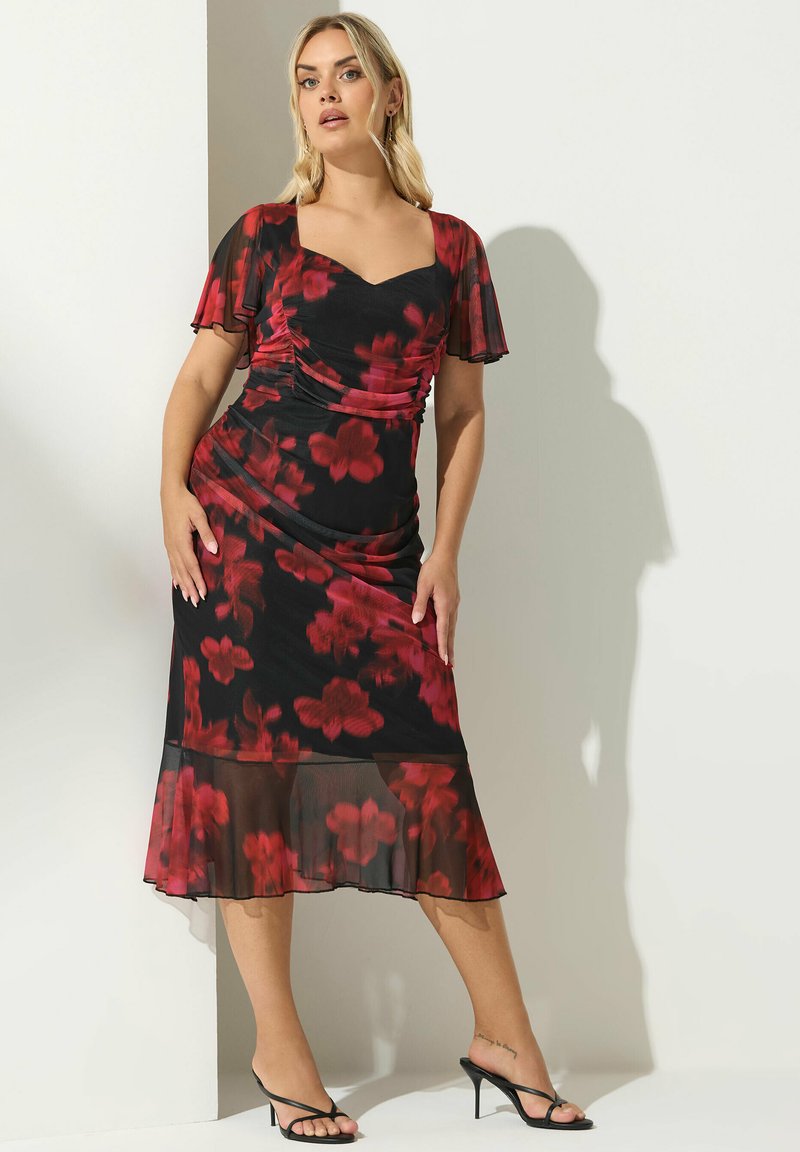 Robe florale noire avec des fleurs rouges, manches courtes en volant, corsage froncé, ourlet transparent et silhouette ajustée. Portée avec des talons noirs à lanières.