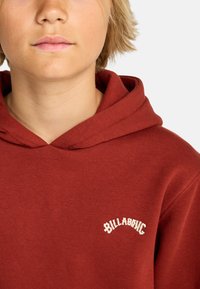 Felpa cappuccio color mogano realizzata in un morbido tessuto, caratterizzata da un logo testurizzato in crema. Design con cappuccio, vestibilità rilassata e polsini a costine.