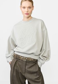 Jasnoszara bluza z haftowanym logo "LM", luźny krój i prążkowany dół. Połączona z brązowymi spodniami w kratę i paskiem w panterkę.