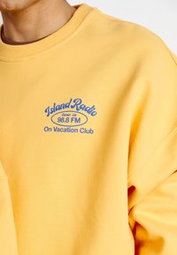 Sárga mikrofonos pulóver, kék grafikával és a "Island Radio 98.8 FM On Vacation Club" felirattal. Bordás nyak és ujj.