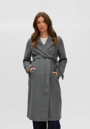 Femme aux longs cheveux bruns portant un manteau en laine gris ceinturé, debout les mains dans les poches devant un fond clair uni.