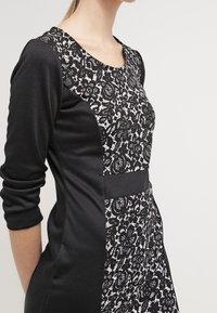 Svart klänning med en figurnära siluett. Har ett texturerat svartvitt spetsmönster på bysten och släta svarta ärmar.