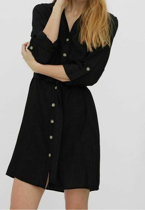 Femme aux longs cheveux blonds portant une robe noire boutonnée sur le devant, manches retroussées et ceinture à la taille, sur un fond clair uni.