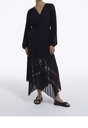 Vestido negro plisado con escote en V y mangas largas, con un panel transparente en la parte inferior y texturas superpuestas. Sandalias planas negras completan el conjunto.