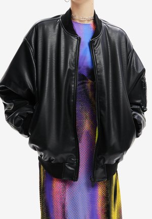 Osoba nosící velkou černou koženou bomber bundu přes barevné vzorované šaty a průhledný řetízkový náhrdelník.