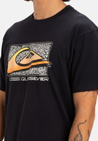 T-shirt nera in cotone con un logo Quiksilver colorato su uno sfondo testurizzato. Il design include elementi grafici e strisce.