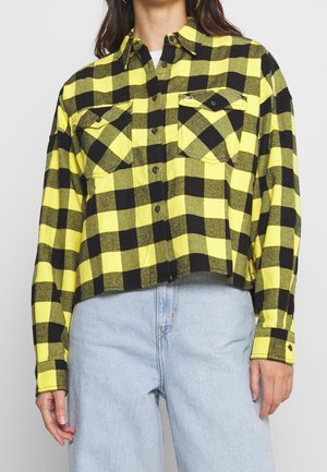 Femme portant une chemise à carreaux jaune et noire boutonnée avec deux poches devant et un jean bleu clair.