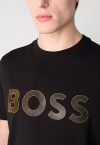Juodas medvilninis marškinėlis su dideliu grafiniu „BOSS“ raštu auksu, turinčiu tekstūruotą, raštuotą dizainą, apvaliu kaklu ir trumpomis rankovėmis.