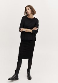 Črn pleteni pulover s tremi četrtinami rokavov v kombinaciji z oprijetim črnim krilom in črnimi gležnarji. Gladek material, minimalističen dizajn.