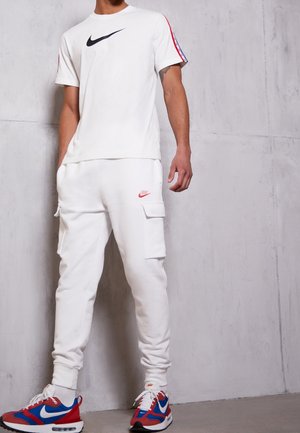 Mann trägt weißes Nike-T-Shirt mit schwarzem Swoosh, weiße Cargo-Jogginghose mit rotem Swoosh und rote, blaue und weiße Nike-Sneakers, steht vor einer grauen Wand.