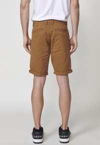 Braune Shorts mit aufgerollten Säumen, Gesäßtaschen und einem Hüftetikett, kombiniert mit schwarzen Sneakers mit weißen Sohlen und Logodetails.