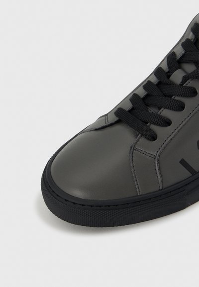 Γκρι δερμάτινο αθλητικό παπούτσι με μαύρη ελαστική σόλα, στρογγυλή μύτη και μαύρα κορδόνια. Διαθέτει ραφές και εκτύπωση λογότυπου στο πλάι.