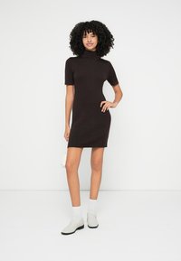 Čierne krátke pletené šaty s rebrovaným vzorom a vysokým golierom. Modelka drží malú bielu kabelku, má na sebe biele ponožky a svetlošedé topánky na nosenie bez šnúrok.