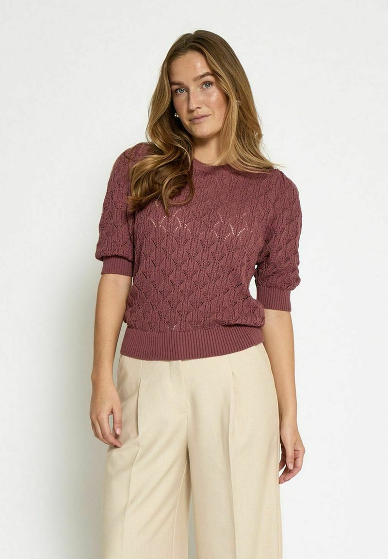 Lilla niidi sviiter, millel on tekstureeritud rombimuster, lühikesed varrukad ja soonikkoes äär. Kombineeritud heledate beežide laia säärega pükstega.