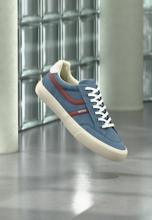 Baskets bleu et blanc avec une bande rouge et le logo « BOSS », flottant à l’intérieur devant un mur en blocs de verre et un sol en béton.