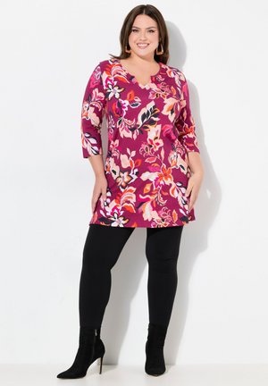 Blumige Tunika in Rosa- und Lilatönen mit Dreiviertelärmeln, kombiniert mit schwarzen Leggings und schwarzen Stiefeletten. Glattes, weiches Material.