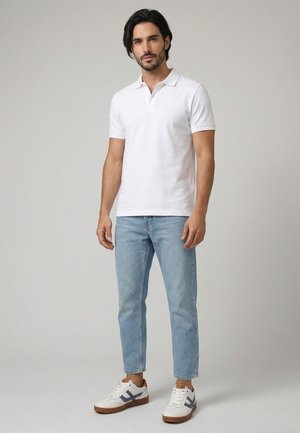 Mann mit dunklem Haar, der ein weißes kurzärmliges Polohemd, hellblaue Jeans und weiße Sneakers mit marineblauen Akzenten trägt, steht vor einem einfachen Hintergrund.