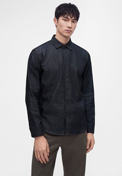 Jeune homme portant une chemise noire à manches longues boutonnée et un pantalon marron, debout devant un fond clair uni.