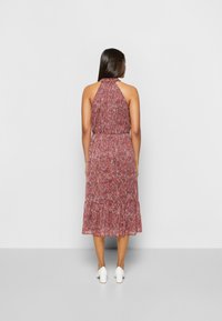 Frau mit langen dunklen Haaren, die ein ärmelloses, halterförmiges, rotes Blumenkleid in Midi-Länge und weiße Blockabsatzschuhe trägt, während sie einer weißen Wand zugewandt ist.
