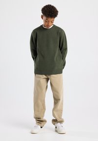 Maglione verde scuro con collo rotondo, abbinato a pantaloni beige a gamba dritta e sneakers bianche. Tessuto testurizzato, vestibilità rilassata.