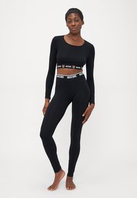 Naine seisab paljajalu, kandes musta pika varrukaga crop-topi ja Moschino logo bändidega legginsit, valge tausta ees.