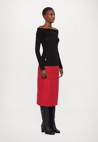 Zwarte off-shoulder top met lange mouwen, gecombineerd met een rode suede midi-rok en zwarte kniehoge haklaarzen met zilveren accenten.