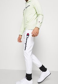 Ljusgrön hoodie med känguruficka och svart broderad "Champion"-logotyp, parat med vita joggingbyxor med röd logotypaccent. Vita sneakers.