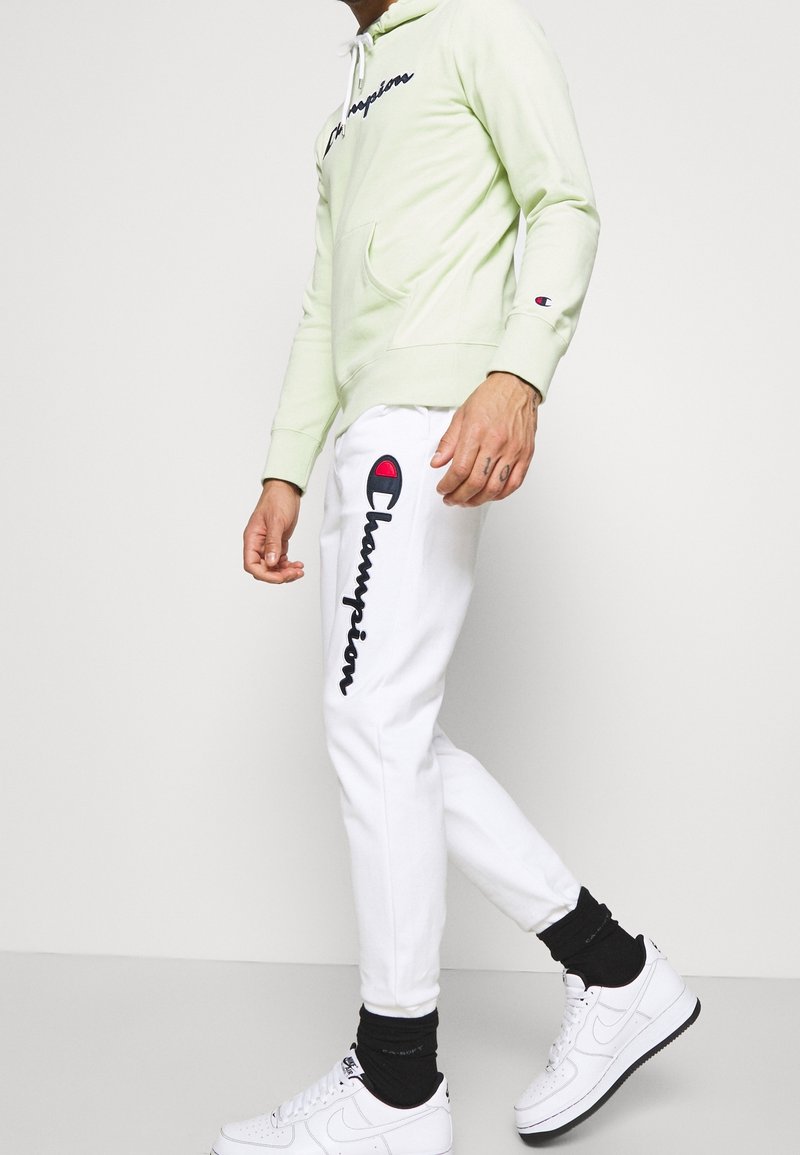 Ljusgrön hoodie med känguruficka och svart broderad "Champion"-logotyp, parat med vita joggingbyxor med röd logotypaccent. Vita sneakers.