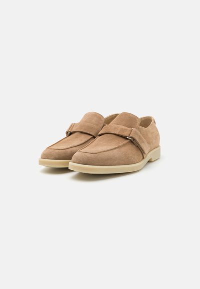 Des chaussures slip-on en suede beige avec un bout arrondi, dotées d'une large sangle et d'un accent circulaire, reposant sur une semelle en caoutchouc de couleur claire.