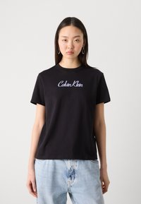 Crna pamučna majica kratkih rukava s okruglim izrezom, na prsima ima ljubičasti izvezeni logo "Calvin Klein".