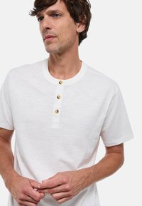 Chemise blanche à manches courtes avec un col rond, dotée d'une patte de boutonnage à trois boutons en accents dorés. Texture douce et coupe décontractée.