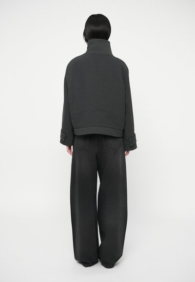 Person med kort, mørkt hår bærer en mørk jakke med høj krave og brede sorte bukser, stående foran en simpel hvid baggrund.