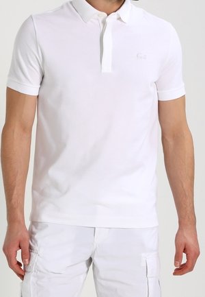 Homme portant un polo blanc uni à manches courtes et un short cargo blanc, debout avec les bras détendus le long du corps.