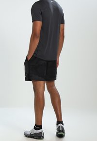 Mężczyzna stoi z rękami w kieszeniach, ubrany w czarne szorty Nike, ciemnoszary T-shirt, czarne skarpetki i szare buty do biegania Nike.