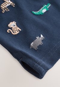 Tissu bleu marine avec de petits animaux brodés, dont un tigre, un crocodile vert, un singe et un rhinocéros gris près d’un bord plié.