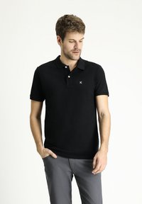 Polo negro con cuello clásico, dos botones frontales, mangas cortas y un pequeño logotipo blanco en el pecho; material de algodón suave.