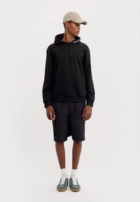 Svart hettegenser med snøring, kengurulomme, kombinert med marineblå shorts. Modellen har på seg en lys beige caps og grønne joggesko med hvite lisser.