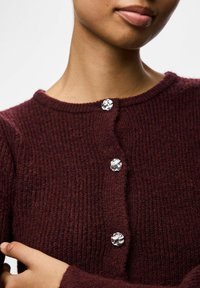 Burgundijas krāsas adīts kardigāns ar ribotu tekstūru, ar skaidriem dekoratīviem pogām priekšpusē un noapaļotu kakla izgriezumu.