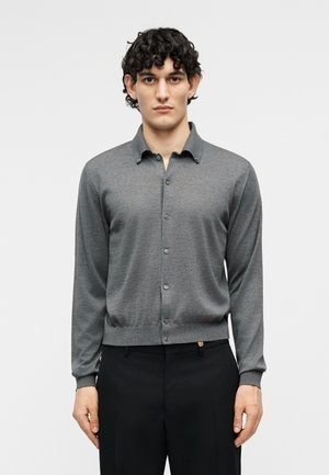 Νέος άνδρας με σκούρα σγουρά μαλλιά φοράει ένα γκρι ζακέτα με κουμπιά και μαύρο παντελόνι, στέκεται με το σώμα στραμμένο προς τα εμπρός μπροστά σε ένα απλό φόντο.