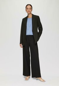 Blazer nero su misura sopra un top azzurro chiaro, abbinato a pantaloni neri a gamba larga e sandali bianchi con cinturini. Tessuto liscio e design strutturato.