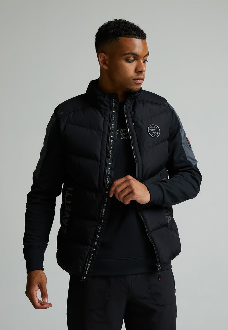 Veste noire matelassée avec une fermeture éclair à l'avant, logo sur la poitrine et accents réfléchissants sur les manches ; elle dispose d'un col haut et de poches latérales.