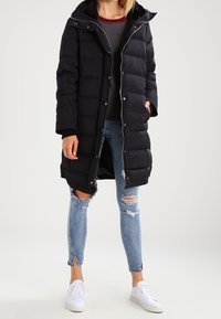 Doudoune noire avec capuche et fermeture éclair à l'avant, présentant un design matelassé et des poches latérales. Portée avec un haut gris et un jean déchiré.