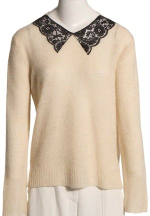 Pull en maille crème à manches longues avec détail de col pointu en dentelle noire, présenté sur un mannequin avec un pantalon blanc.