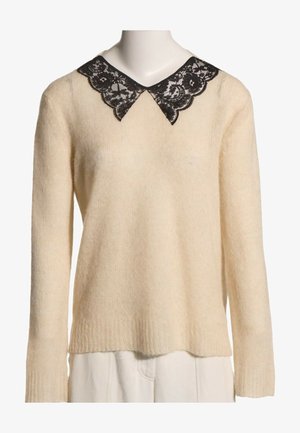 Pull en maille crème à manches longues avec détail de col pointu en dentelle noire, présenté sur un mannequin avec un pantalon blanc.