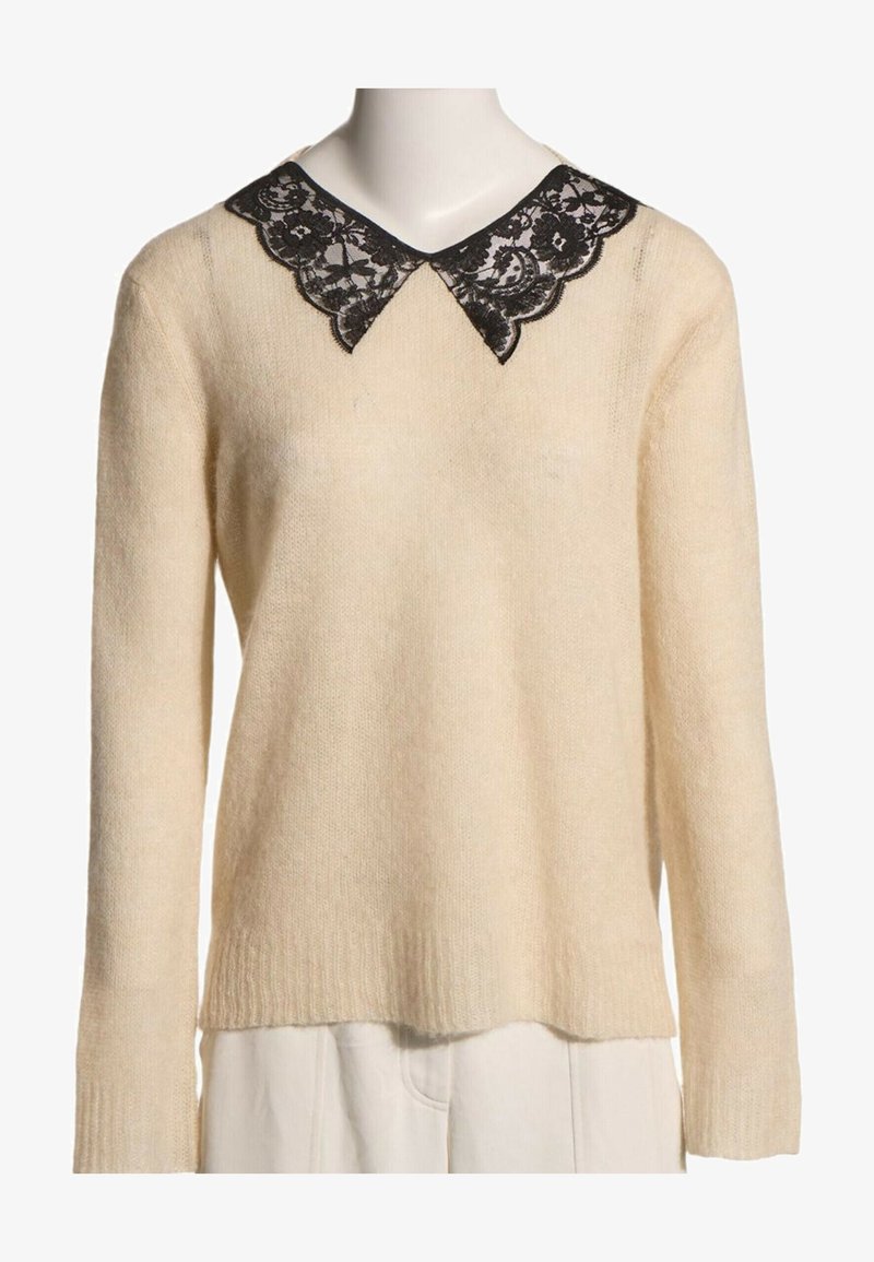 Pull en maille crème à manches longues avec détail de col pointu en dentelle noire, présenté sur un mannequin avec un pantalon blanc.