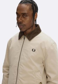 Giacca beige con colletto in velluto a coste marrone, dotata di cerniera frontale, due tasche laterali e un logo nero sul petto a sinistra.