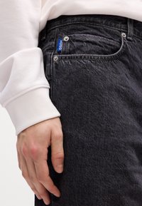 Hånd hvilende mod sorte denimjeans med synlig "HUGO" mærkeetiket, bar med en hvid langærmet tøjmuffe.