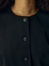 Prenda de tela negra con cuello redondo y cinco botones negros texturizados en la parte delantera. Textura suave con un ajuste entallado.