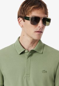 Un giovane indossa una polo verde chiaro e occhiali da sole grandi e alla moda, mostrando un'espressione neutra su uno sfondo bianco.