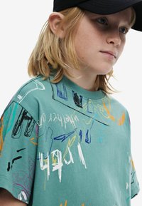 Bavlnené tričko v tyrkysovej farbe s grafickými motívmi v štýle graffiti v rôznych farbách. Má okrúhly výstrih a krátke rukávy, s voľným strihom a jemnou textúrou.